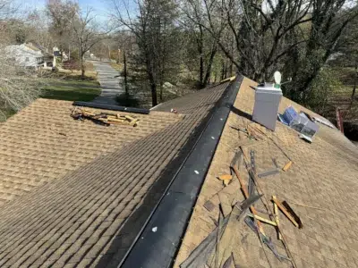 Residential roof vent installation on a tan shingle roof in King of Prussia, PA, featuring a chimney with a missing or damaged chimney cap. Roofing tools, vent materials, and debris are scattered on the roof, with a neighborhood and trees visible in the background.